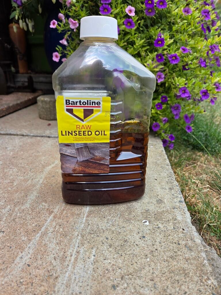 A plastic bottle of Bartoline raw linseed oil with a yellow label, sitting outdoors on a stone patio with purple flowers in the background.