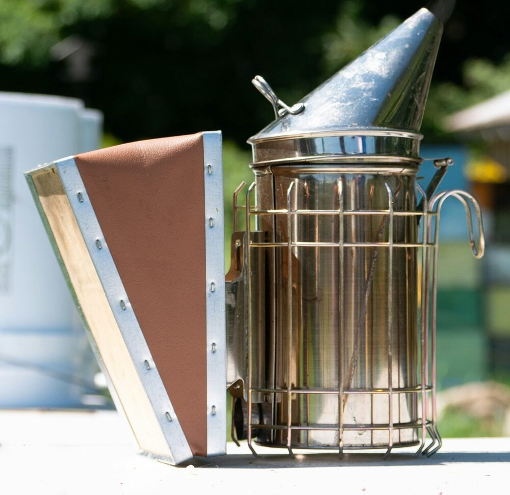Stainless steel bee smoker with a protective heat guard and leather bellows emitting a faint trail of smoke.