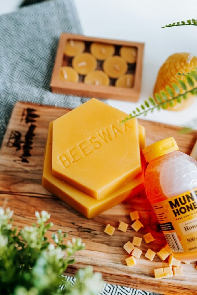Hexagonal yellow beeswax block displayed with a bottle of honey, wax pellets, and beeswax tealight candles on a wooden board